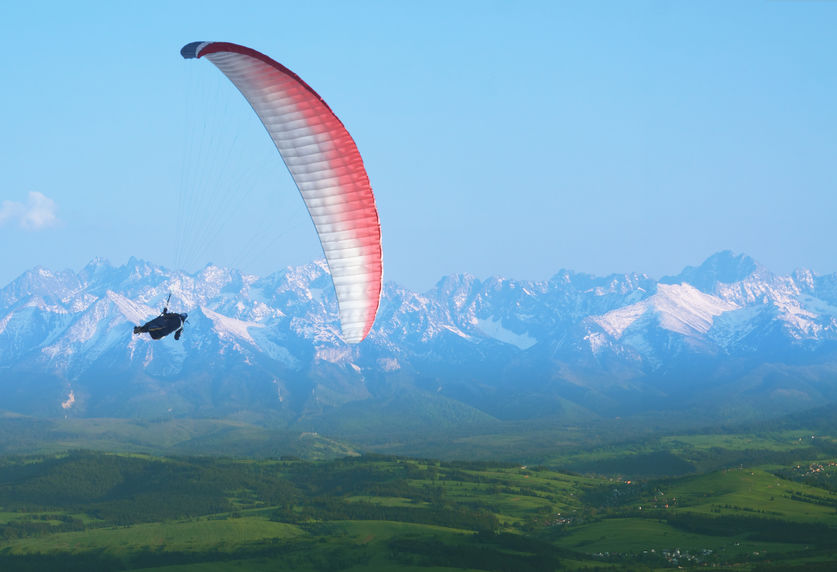 Parapentiste en vol au-dessus d’une vallée verdoyante, avec en arrière-plan une chaîne de montagnes enneigées sous un ciel bleu clair.
