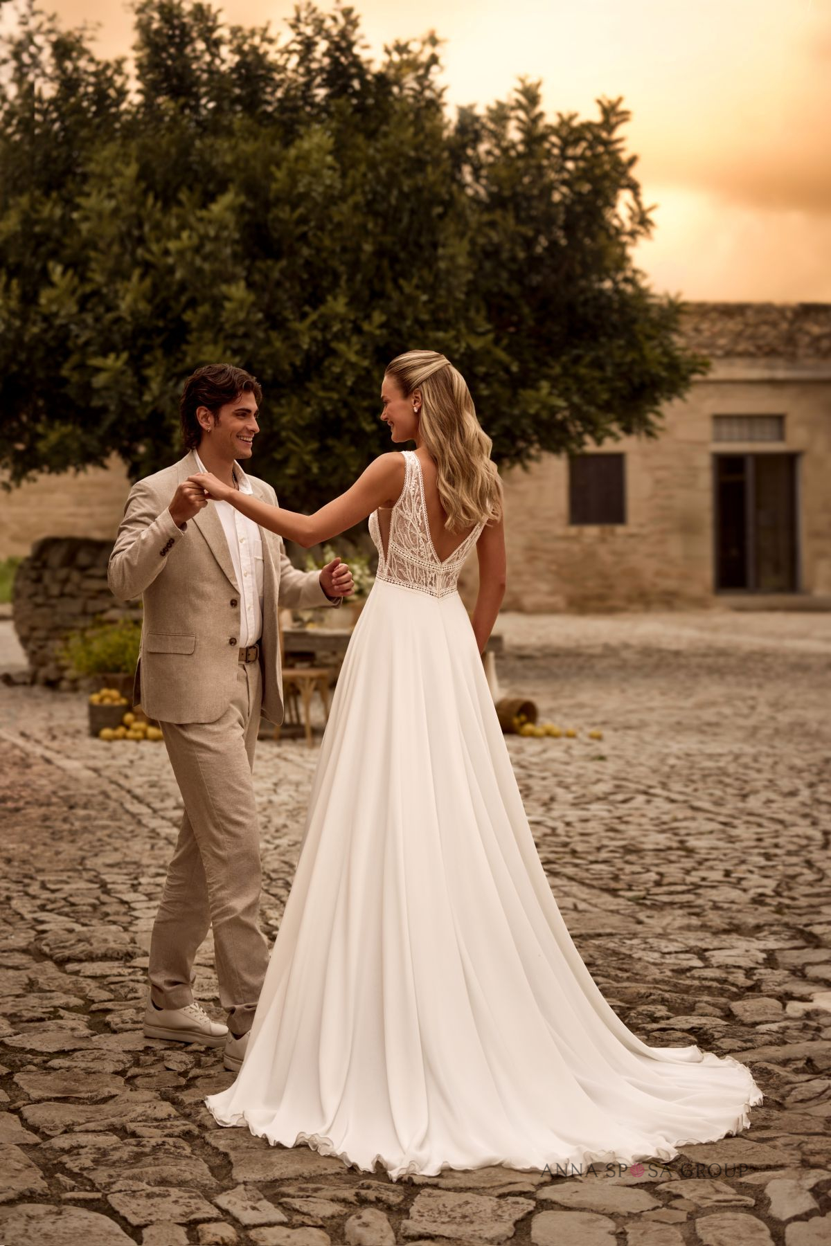 Couple dans une cour en pierre au coucher du soleil, la mariée portant une robe fluide au dos nu en dentelle, style bohème chic.