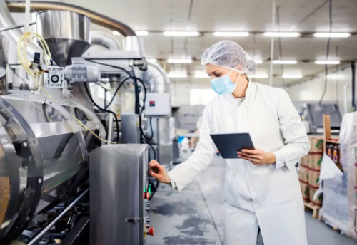 Femme ingénieure en blouse blanche, masque et charlotte, inspectant une machine industrielle dans une usine agroalimentaire à l’aide d’une tablette numérique.
